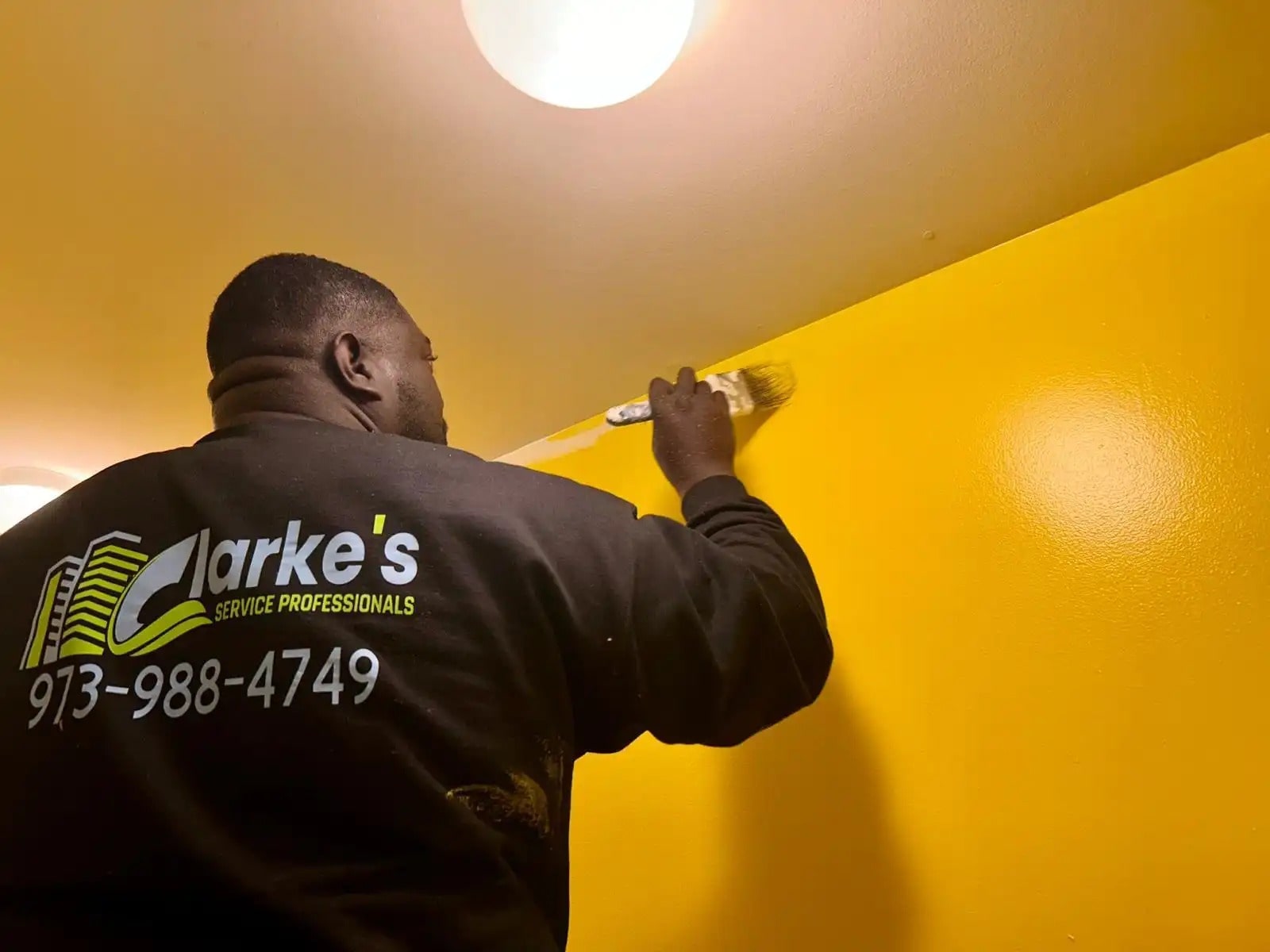 Man painting a bright yellow wall with a brush, wearing a "Clarke's Service Professionals" shirt. Call 973-988-4749.