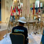 Clarke's Service Professionals Technician assembles chandelier beneath flags and stage in grand hall