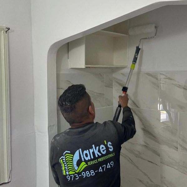 Man painting bathroom with roller, Clarke's Service Professionals logo on shirt, phone 973-988-4749.