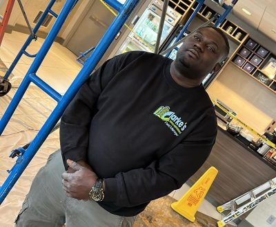 c7 Construction worker in black "Clarke's Field Manager" sweatshirt stands near scaffolding.