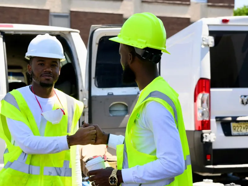 dfsdfsdfsdf-scaled (1)-optimized Two construction workers shake hands, wearing safety vests and hard hats near work vans.